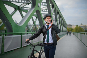Businessman commuter on the way to work, pushing bike on bridge and calling on mobile phone, sustainable lifestyle concept.