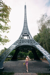 A stylish slender smiling girl with blond long hair stands with a bouquet of wild flowers in her hands, near a copy of the Eiffel Tower in Ukraine. Dressed in a bright summer skirt and a sexy top.