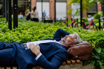 Mature businessman relaxing on bench in city park during break at work, work-life balance concept.