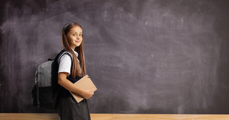 Schoolgirl with a backpack and book standing in front of a blank blackboard