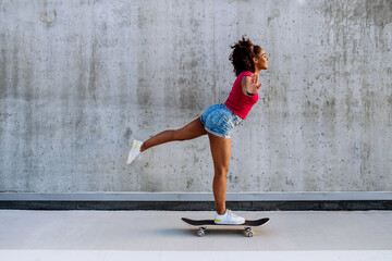 Multiracial teenage girl riding a skateboard in front of concrete wall, standing in one leg, balancing. Side view. - Powered by Adobe