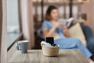 digital detox and leisure concept - close up of gadgets in basket on table and woman reading book at home - obrazy, fototapety, plakaty