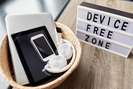digital detox and technology concept - close up of device free zone words on light box and different gadgets in wicker basket on table at home