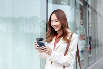 Fototapeta na wymiar Beautiful asian female businesswoman use smartphone hold paper cup of hot drink, Walk enjoy smiling while doing commuting in the modern city near office building outside