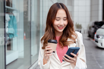 Beautiful asian female businesswoman use smartphone hold paper cup of hot drink, Walk enjoy smiling while doing commuting in the modern city near office building outside