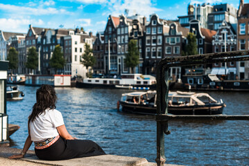 Mujer joven descansando sentada a orillas de un canal en un día soleado de verano en la ciudad de Ámsterdam. Un gran barco navegando por el canal frente a varios edificios típicos holandeses. - obrazy, fototapety, plakaty