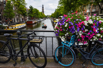 Fototapeta na wymiar Dos bicicletas aparcadas en un puente de Ámsterdam adornado con flores moradas y rosas. En el canal varios barcos y barcas de distintos tamaños. Al fondo, la torre de la iglesia Westerkerk.