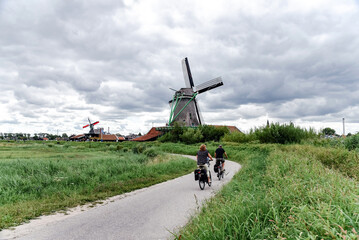 Paisaje del pueblo de Zaanse Schans, en Países Bajos. Dos personas montando en bicicleta con molinos de viento tradicionales de fondo en un día de verano nublado.  © AliciaFdez