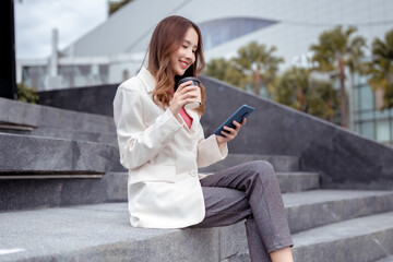 Beautiful asian female businesswoman use smartphone hold paper cup of hot drink, Walk enjoy smiling while doing commuting in the modern city near office building outside