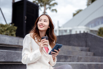 Beautiful asian female businesswoman use smartphone hold paper cup of hot drink, Walk enjoy smiling while doing commuting in the modern city near office building outside