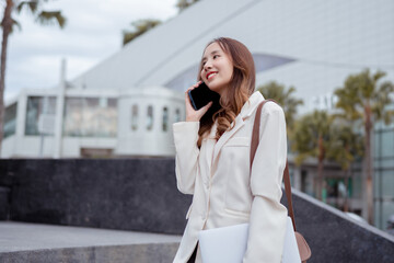 Fototapeta premium Beautiful asian female businesswoman use smartphone and holding laptop computer, Walk enjoy smiling while doing commuting in the modern city near office building outside