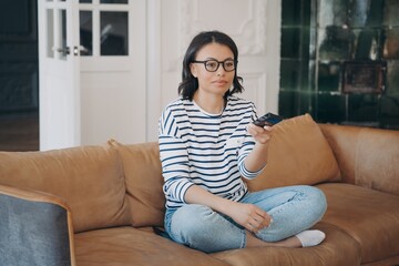 Obraz premium Female wearing glasses changing television channels, watching TV, resting, sitting on sofa at home