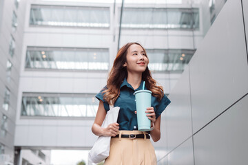 Smiling asian businesswoman hold reusable eco-friendly ecological cup and paper bag while commuting in city in the morning