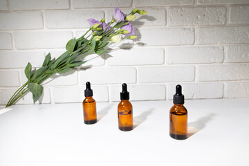 A group of amber bottle on a white table with face serum products near purple eustoma. Glass bottle with self care product. Front view