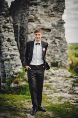 The attractive groom with bow tie in a suit with boutonniere or buttonhole on jacket, is stands on the background stony, rocky wall. Wedding ceremony in country.