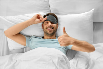 people, bedtime and rest concept - happy smiling man in sleeping eye mask lying in bed and showing...