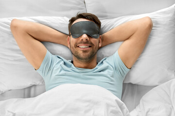 people, bedtime and rest concept - happy smiling man in sleeping eye mask lying in bed, top view