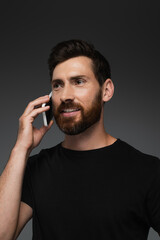 joyful and bearded man in black t-shirt talking on smartphone isolated on grey.