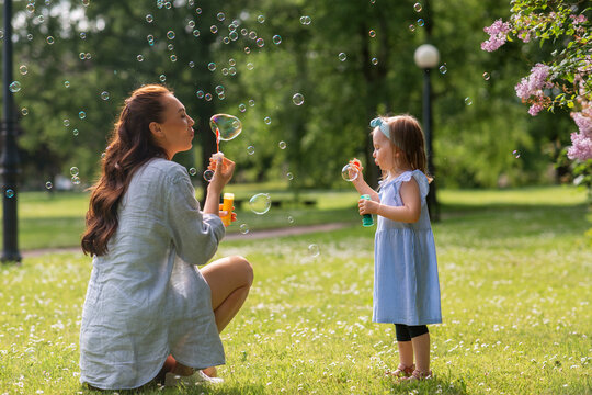 family, motherhood and people concept - happy mother with little daughter blowing soap bubbles at summer park or garden - Powered by Adobe
