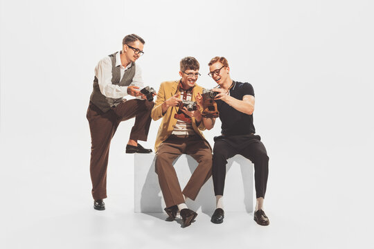 Portrait of three stylish young men in trendy suits sitting, looking at retro camera photos isolated over white studio background