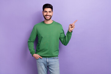 Photo of handsome good mood man wear green long sleeve directing at empty space arm in pocket isolated on purple color background