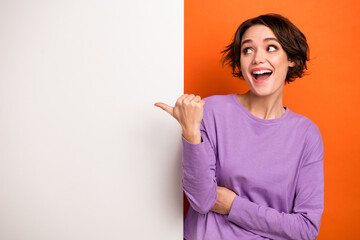 Photo of adorable excited girl dressed purple pullover pointing thumb white placard empty space isolated orange color background