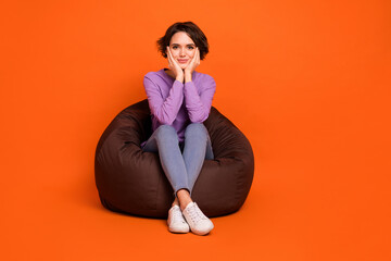Full length photo of shiny dreamy lady wear violet sweatshirt sitting bean bag arms cheeks isolated orange color background