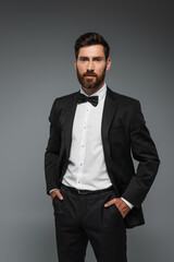 bearded man in elegant tuxedo with bow tie posing with hands in pockets isolated on grey.