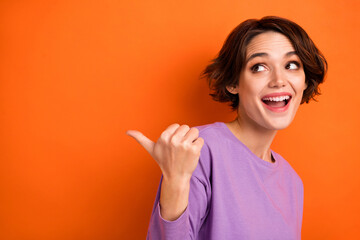 Photo of funny excited girl dressed purple pullover pointing thumb back looking empty space isolated orange color background