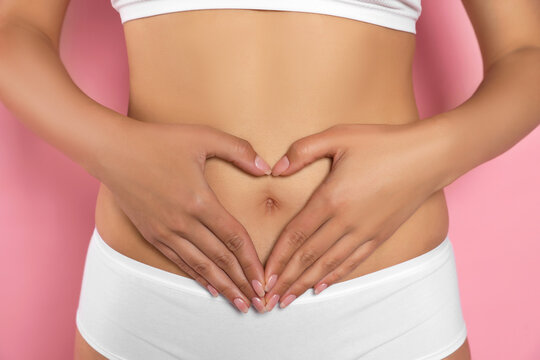 Woman in underwear making heart with hands on her belly against pink background, closeup. Healthy stomach