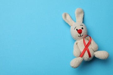 Cute knitted toy bunny with red ribbon on blue background, top view and space for text. AIDS disease awareness