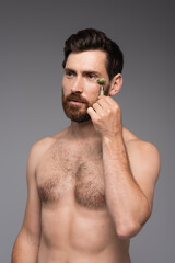 Fototapeta premium shirtless man with beard using jade roller while massaging face isolated on grey.