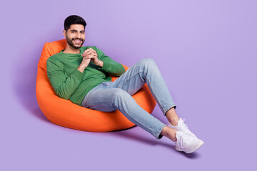 Full size photo of young latin man sit comfy beanbag hold hands toothy smile wear stylish green clothes isolated on purple color background