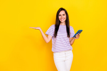 Photo of positive cute lady wear stylish striped outfit open arm hold telephone demonstrate empty space isolated on yellow color background