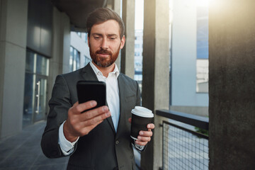 Attractive businessman holding phone standing near office building during break