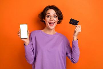 Photo of excited girl demonstrate empty space display debit card isolated on orange color background