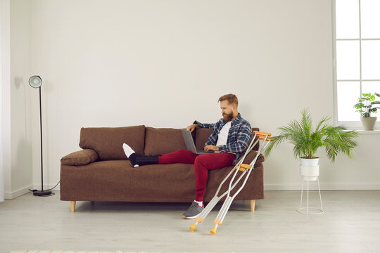 Man With Leg Injury Uses Laptop To Work From Home, Watch Movies, Search For Information On Internet And Order Delivery. Man Sitting On Sofa In Living Room Near Crutches And Looking At Laptop Screen.