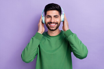 Photo of handsome toothy beaming man dressed green long sleeve hands on headphones listening music isolated on purple color background