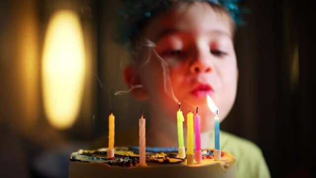 Close-up of charming caucasian little boy making wish and blowing candles on birthday cake. Portrait of happy pretty child celebrating birthday indoors at home. Child blowing out candles on cake