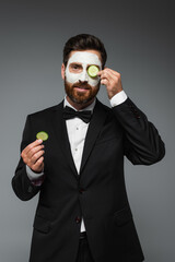 bearded man in suit with clay mask on face holding slices of cucumber isolated on grey.