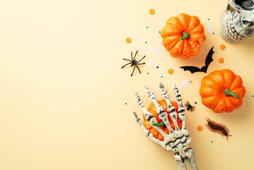 Halloween creepy decorations concept. Top view photo of skull skeleton hand holding pumpkins bat silhouette spider centipede and confetti on isolated beige background with copyspace