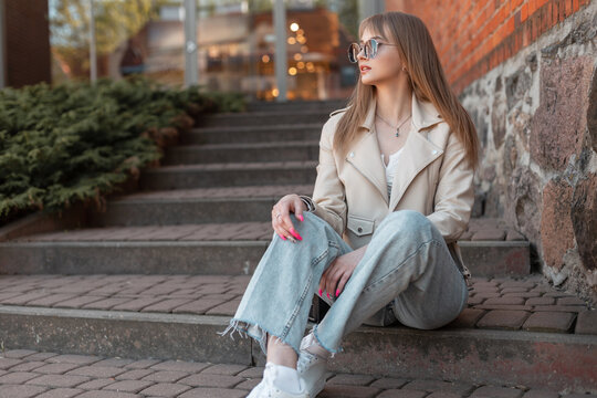 Stylish beautiful young woman with sunglasses in fashionable casual clothes with a leather jacket, top and jeans with white sneakers sits on the steps near a vintage brick building shopping mall. - Powered by Adobe