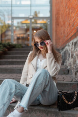 Fashionable adorable stylish young woman wears sunglasses in leather jacket and vintage blue jeans with bag sits on the steps near a shopping mall. Trendy female handbag