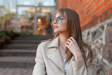 Urban female portrait of a beautiful young fashion woman with stylish vintage round sunglasses in a fashionable leather jacket and T-shirt sits on the steps near a vintage brick building mall