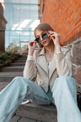 Cool beautiful young stylish woman hipster in fashionable clothes with a leather jacket and jeans sitting on the steps and wearing round vintage sunglasses near the mall in the city. Fashion style