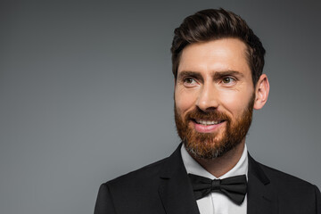 cheerful and bearded man in black tuxedo with bow tie isolated on grey.