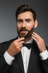 portrait of happy man in tuxedo brushing beard with comb isolated on grey.