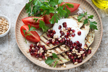 Grilled slices of aubergine served with pomegranate seeds, pine nuts, white yogurt, red tomatoes and parsley on a beige plate, middle closeup