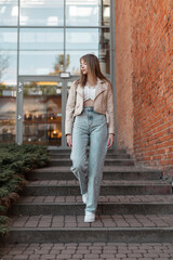Trendy beautiful young woman in fashionable casual wear outfit with a leather jacket with bell-bottom high-waisted jeans and white sneakers walks near a red brick store building