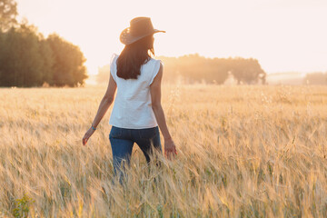 Fototapeta na wymiar Woman farmer in cowboy hat walking with hands on ears at agricultural rye field on sunset.